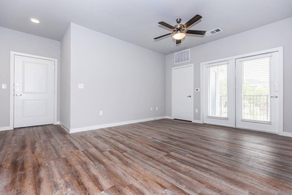 A spacious, unfurnished room featuring light gray walls, a ceiling fan, and brown hardwood flooring. There are two sets of double doors leading to a balcony, a white door on the right, and a front door on the left. Natural light fills the space, creating an inviting atmosphere.