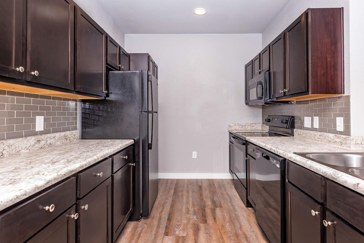 A modern kitchen featuring dark wooden cabinets, a black refrigerator, and stainless steel appliances. The countertops are speckled gray, with a backsplash of gray tiles. The space has wooden flooring and a neutral-colored wall, giving it a contemporary and clean look.