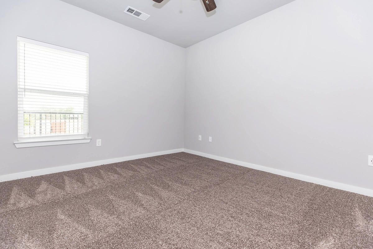 A vacant room with gray walls and a light-colored carpet. There’s a window with white blinds on one wall, allowing natural light to enter. The ceiling features a ceiling fan, and the room is free of furniture, giving it an open and spacious appearance.