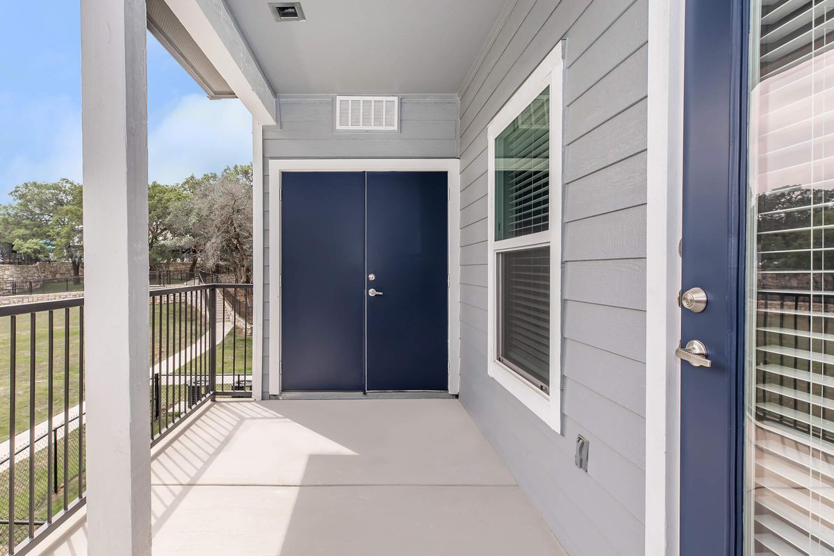 A view of a modern porch outside a home, featuring a blue front door, light gray siding, and large windows. The porch has a concrete floor and a railing, with green grass visible beyond the entrance. The scene is bright and inviting, suggesting a comfortable living space.