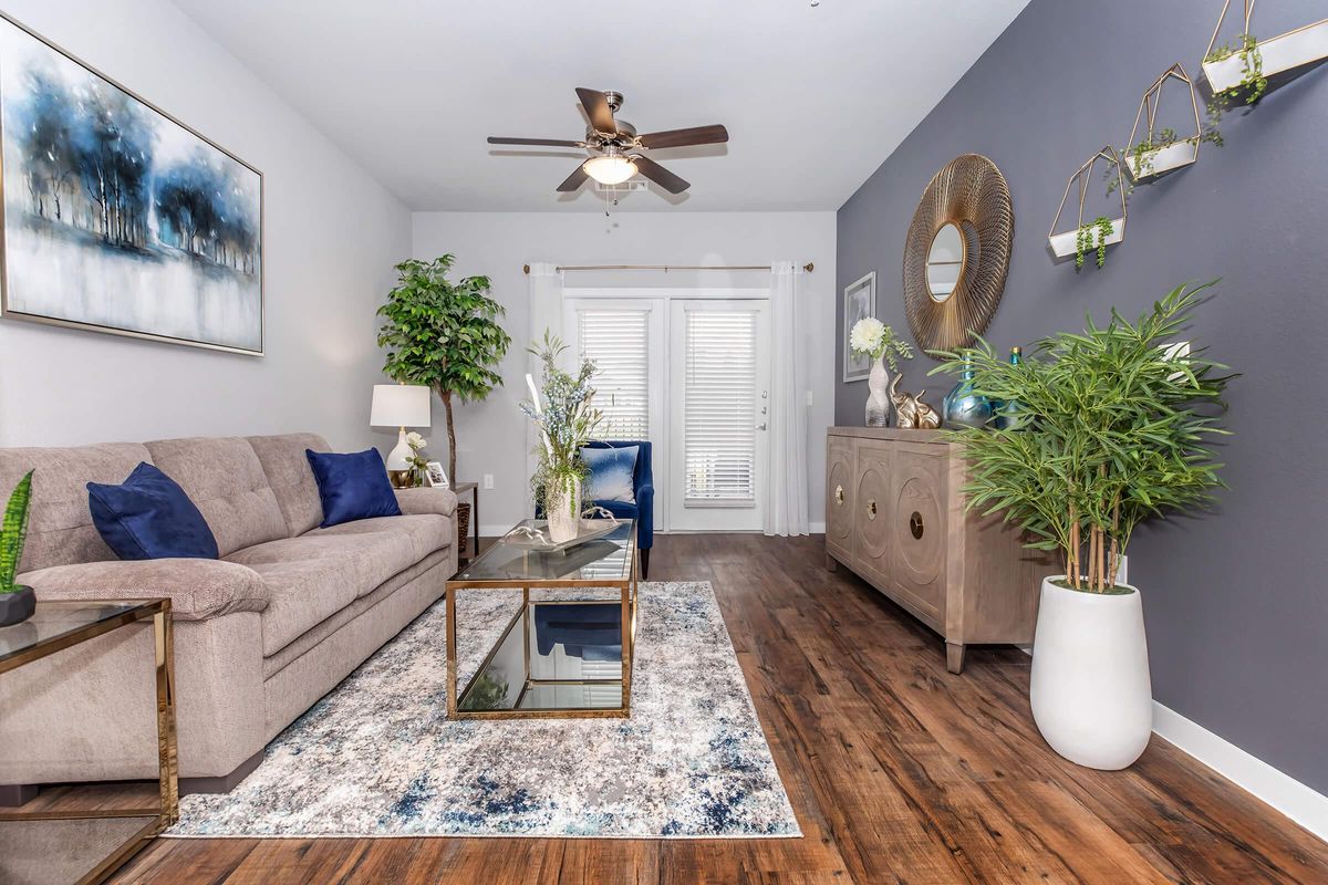 A modern living room featuring a light gray sofa, a glass coffee table, and a decorative area rug. The walls are painted in shades of gray and white. There's a ceiling fan, potted plants, and stylish decor items on a sideboard. Natural light streams through the window leading to a patio.