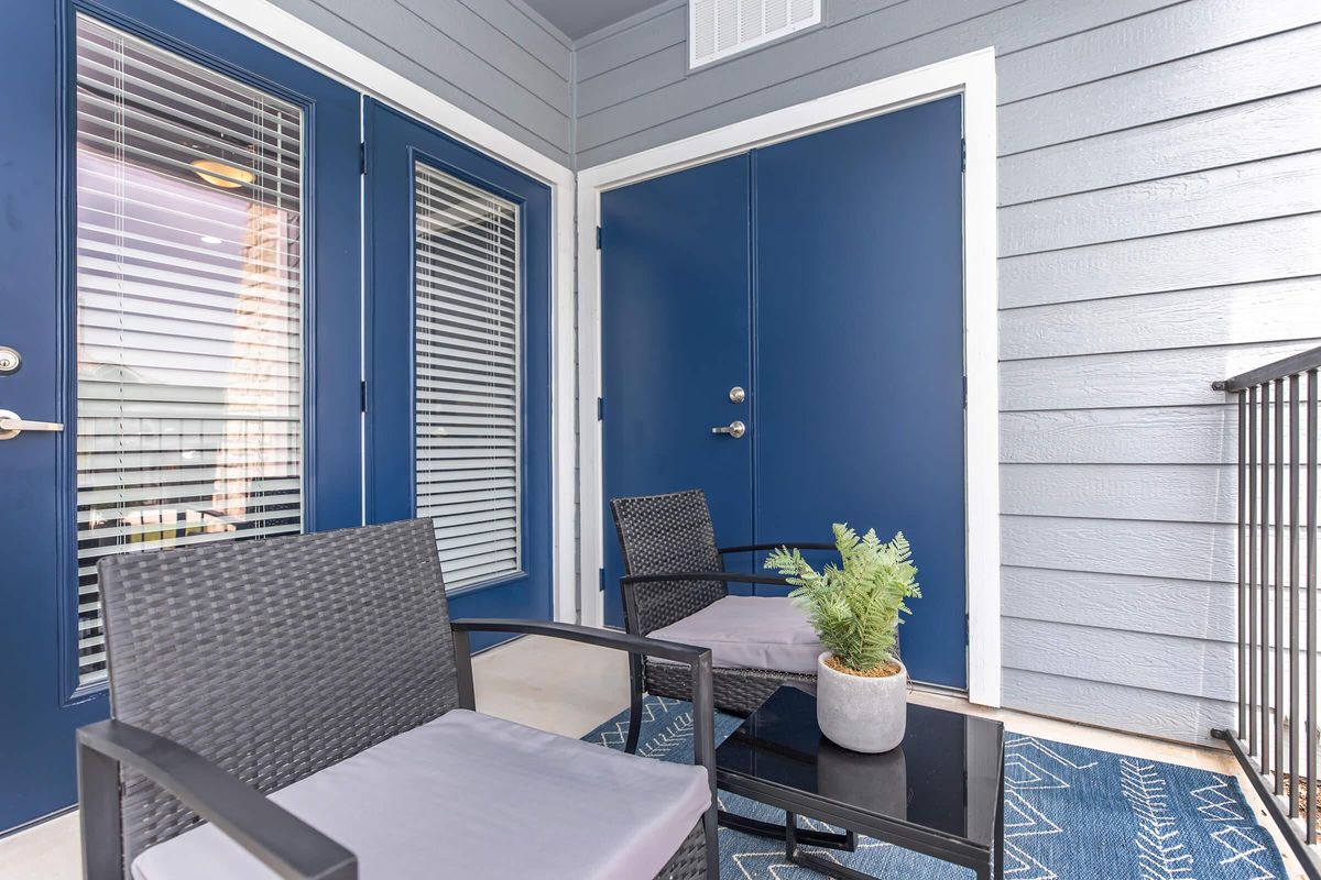 A cozy outdoor space featuring two black wicker chairs with gray cushions, a small black table, and a potted plant on the table. The background includes a pair of blue doors with glass panels and vertical blinds, set against a light gray wood-paneled wall.