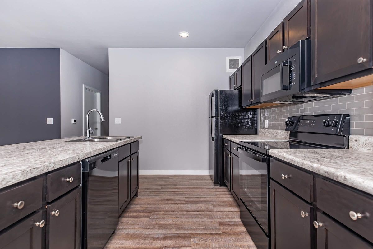 Modern kitchen featuring dark cabinetry, granite countertops, and stainless steel appliances. The space includes a black refrigerator, microwave, and electric stove, with a sink integrated into the kitchen island. Light gray walls and wood-look flooring enhance the contemporary design.
