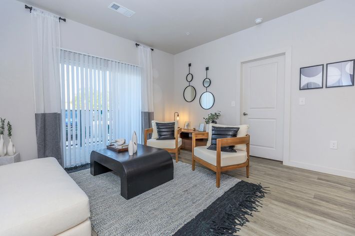 A modern living room featuring two stylish wooden chairs with white upholstery, a black coffee table, and a textured area rug. The space includes large windows with sheer curtains and a sliding door, along with decorative mirrors and artwork on the walls, creating a cozy and inviting atmosphere.