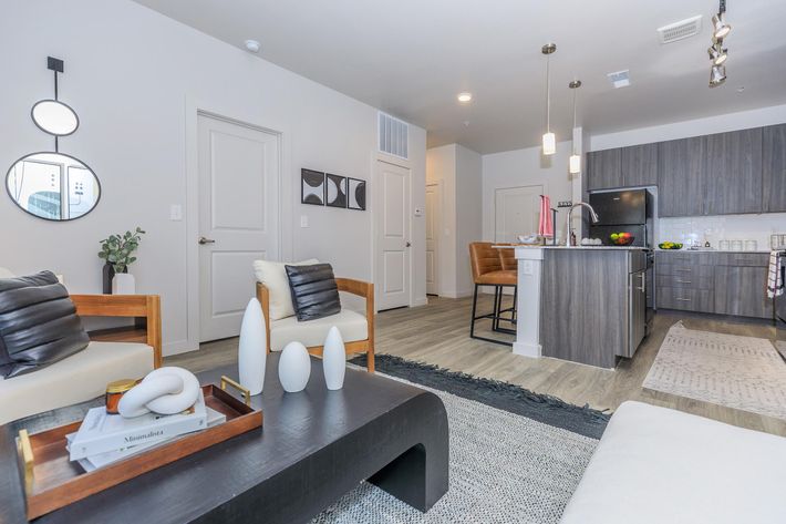 A modern living space featuring a cozy seating area with two wooden chairs and a white sofa. A dark coffee table holds decorative items. In the background, a kitchen island with bar stools is visible, alongside stainless steel appliances. The decor is minimalistic and stylish, with a neutral color palette.