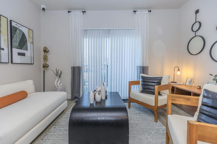 A modern living room featuring a white sofa with an orange accent pillow, two wooden chairs with black cushions, a black coffee table, and decorative vases. Light-colored curtains cover a window, and abstract artwork adorns the walls, creating a cozy and stylish atmosphere.