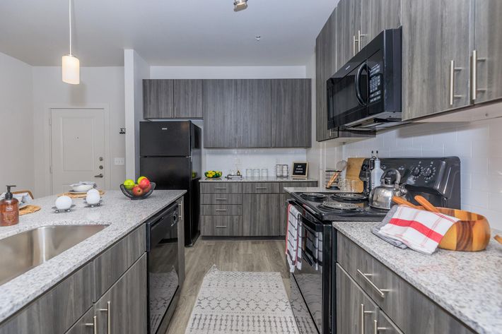 Modern kitchen with dark wood cabinetry, a gray speckled countertop, and stainless steel appliances. The layout includes a black refrigerator, microwave, and oven. A small island features a wooden bowl with fruits, and there are decorative elements like dish soap and towels. Light enters through a nearby window.