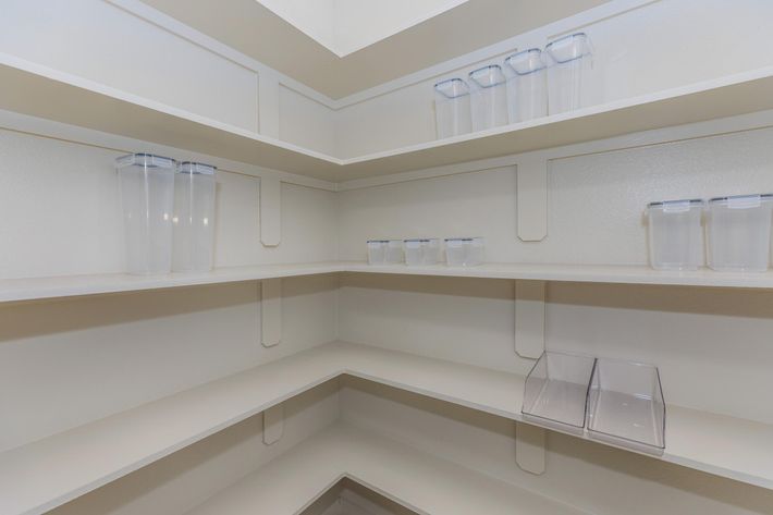 A clean and organized pantry with light-colored shelves. Four transparent storage containers are neatly arranged on the top shelf, and several smaller clear bins are placed on the middle shelves, creating a tidy and minimalistic appearance. The overall atmosphere is bright and spacious.