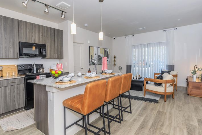 A modern kitchen and living area featuring a countertop with bar stools, black appliances, and decorative table settings. Light-colored walls, stylish furnishings, and large windows offer a bright, inviting atmosphere. A cozy seating area is visible in the background, enhancing the contemporary decor.