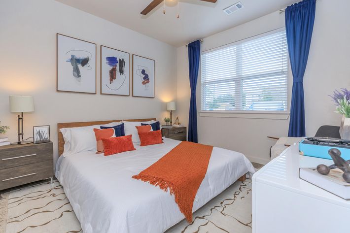 A cozy, modern bedroom featuring a neatly made bed with white linens and orange accent pillows. Two bedside lamps flank the bed, and there are three framed artworks on the wall above. The room has a large window with white blinds and blue curtains, allowing natural light to brighten the space, complemented by a decorative rug.