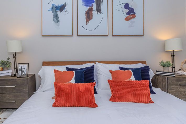 A cozy bedroom featuring a neatly arranged bed with white linens and colorful pillows in shades of orange and navy. Flanking the bed are two wooden nightstands with lamps, and above the bed are three abstract framed artworks. The room has a warm and inviting atmosphere.