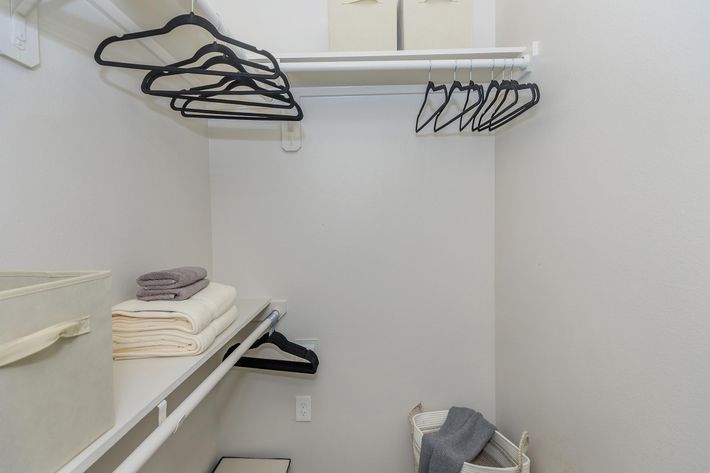 A neatly organized closet with a white wall. It features a rod with multiple black hangers, rolled towels, and storage bins. A shelf holds folded blankets, while a laundry basket rests on the floor, contributing to a clean and tidy appearance.