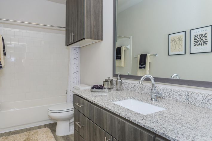 A modern bathroom featuring a light-colored tub and shower combination with a glass sliding door. The countertop is made of speckled granite, adorned with a small decorative tray and a soap dispenser. There are dark wooden cabinets, and two framed black-and-white artworks on the wall. A large mirror reflects the space.