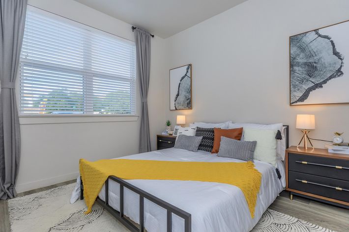 A cozy bedroom featuring a neatly made bed with decorative pillows and a yellow throw blanket. Two framed artworks hang on the light-colored walls, and a window with white blinds allows natural light to brighten the space. A stylish nightstand with lamps and a small plant adds to the modern decor.