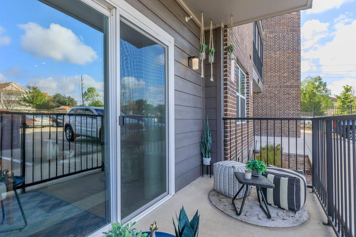 A cozy balcony view featuring a glass door, a small table with decorative pillows, and potted plants. The balcony is enclosed with a black railing and overlooks a parking area with trees in the background. Bright blue skies and fluffy clouds enhance the serene atmosphere.