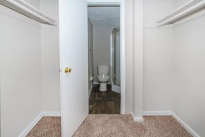 A clean, simple bathroom with a toilet visible through an open door. The space features beige walls and light-colored carpet, with a small corner shower visible. There are two empty closet spaces on either side of the entryway, adding to the minimalistic layout.