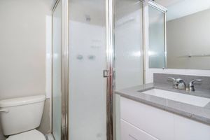 A modern bathroom featuring a glass shower stall, a toilet, and a sleek sink with a gray countertop. The walls are light-colored, and the shower area has a frosted glass door for privacy. Bright lighting enhances the clean, minimalist design.