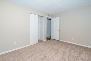Empty room with light beige walls and carpeted floor. A small closet with sliding doors is on the left, and a doorway leads to another room on the right. The space is well-lit and minimalistic, creating a neutral and inviting atmosphere.