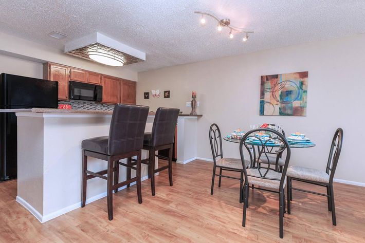 A modern dining area featuring a table set for four, with decorative plates and glasses. Nearby is a kitchen bar with high stools. The kitchen has wooden cabinetry and black appliances. The walls are painted in neutral tones, complemented by a colorful abstract painting. The flooring is light wood.