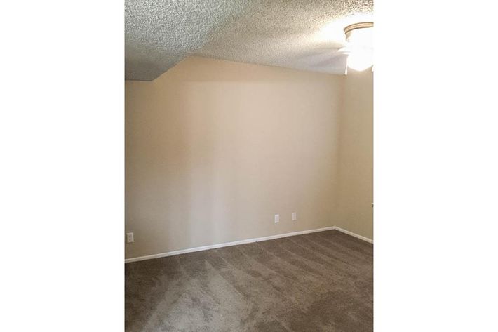 Empty room with a light beige wall and a light-colored carpet. A ceiling fan is visible, and there are no furnishings or decorations. The room is well-lit and appears spacious. A sloped ceiling is present on one side, creating a unique architectural feature.