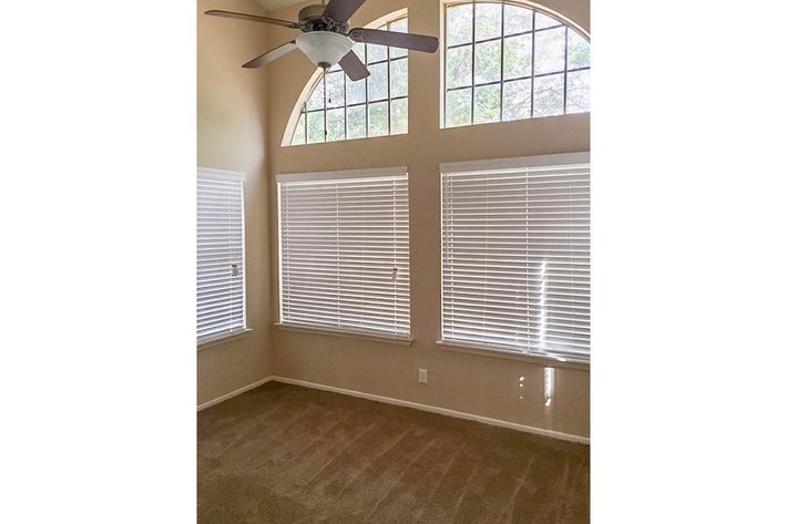 A bright, empty room with light beige walls and a tan carpet. It features large arched windows with white blinds, allowing natural light to illuminate the space. A ceiling fan is installed in the center of the room, adding a touch of modern design. The overall atmosphere is spacious and inviting.