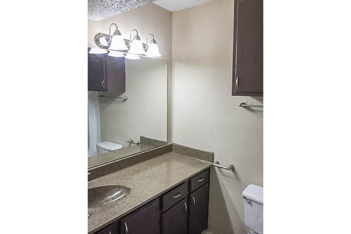 A clean bathroom featuring a light fixture with four bulbs above a mirror. The countertop is made of a speckled stone material, with a sink and cabinet below. There's a towel bar on the wall and a toilet visible to the right. The walls are painted a neutral color.