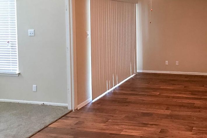 A room featuring two different flooring types: carpet on the left and wooden planks on the right. There are white vertical blinds covering a window or door, allowing soft light to filter in, and light beige walls. The space appears open and well-lit.