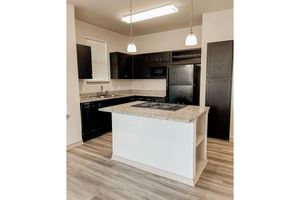 Modern kitchen featuring dark cabinetry, a granite countertop with an island cooktop, and stainless steel appliances. The room is well-lit with pendant lights hanging above the island and has a light-colored wood floor. A window with vertical blinds adds natural light to the space.