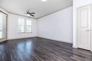 Spacious and empty room with dark wood-like flooring, large windows with blinds allowing natural light, a ceiling fan, and a single door on the right. The walls are painted white, contributing to a bright and airy atmosphere, suitable for various interior design possibilities.
