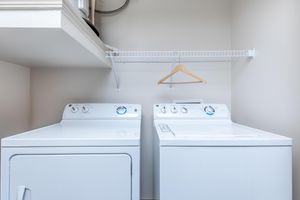 A clean laundry room featuring a white washing machine and a dryer side by side. Above, there's a wire shelf with a wooden hanger. The walls are a light color, creating a bright and tidy space for laundry tasks.