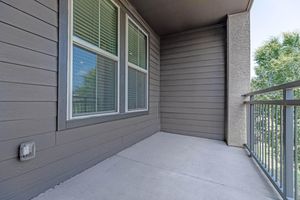 A clean and empty balcony with a gray wooden exterior and large windows. The concrete floor is smooth, and the railing is metal. There are trees visible outside, indicating a quiet, outdoor space.