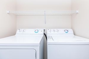 A clean laundry room featuring a white washing machine and a dryer side by side. Above them, a wire shelf is mounted on a light-colored wall, providing space for storage. The appliances have control dials and knobs visible, indicating their settings.