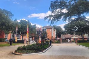 Entrance to a residential complex featuring a landscaped area with flowers and decorative flags. The sky is partly cloudy, and several trees are visible in the surrounding area.