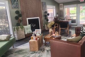 A modern, stylish living room featuring a mix of seating options including a green sofa, a brown leather chair, and wooden coffee tables. The interior is decorated with greenery, abstract wall art, and a fireplace. Large windows allow natural light to fill the space, creating a bright and inviting atmosphere.