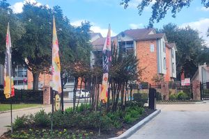 A view of an apartment community featuring several colorful flags, landscaped greenery with flowers and bushes, and a gated entrance. The building is made of red brick, and there are trees in the background under a clear blue sky.