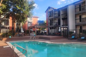 A view of a swimming pool surrounded by wooden decking and lounge chairs, with apartment buildings in the background and green trees providing shade. The scene is well-lit, showcasing a clear blue sky and a relaxed atmosphere.