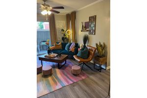 A cozy living room featuring a green sofa adorned with colorful pillows, a stylish brown armchair, and a wooden coffee table. There are decorative plants, a colorful area rug, and a fan on the ceiling. Natural light streams in through a window, creating a warm and inviting atmosphere.