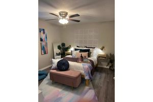 A modern bedroom featuring a neatly made bed with colorful pillows and a light comforter. There is a pink bench at the foot of the bed and a patterned wall behind it. The room includes decorative plants, artwork on the walls, and a mirror, all illuminated by a ceiling fan light.