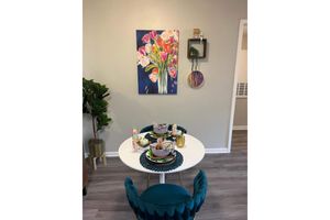 A cozy dining area featuring a round white table set with bowls and drinks, surrounded by two teal chairs. A vibrant floral painting hangs on the wall above the table, with a small decorative shelf holding a circular art piece. A potted plant adds greenery to the space, enhancing the inviting atmosphere.