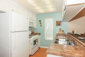 Bright and modern kitchen featuring white cabinetry, a large refrigerator, and a gas stove. The countertop is made of a light brown material, and there are two sinks. A window lets in natural light, and the walls are painted a soft turquoise. Decorative items are placed on the countertops.