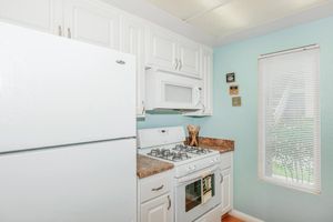 A bright kitchen featuring white cabinetry, a white refrigerator, and a gas stove. A microwave is mounted above the stove. The countertop is a light-colored stone, and there are utensils in a holder. A window with blinds lets in natural light. The walls are painted a soft blue, creating a fresh atmosphere.