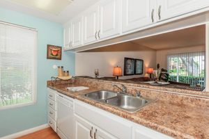 A modern kitchen featuring a double sink, light-colored cabinets, and a granite countertop. There is a window with blinds allowing natural light in, and a cozy living area visible through an opening. Decorative items are placed on the countertop, adding to the inviting atmosphere.