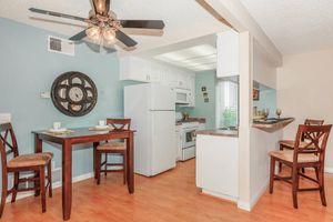 A cozy kitchen and dining area featuring a small wooden dining table with two place settings, a nearby breakfast bar with two stools, white cabinetry, a refrigerator, and an oven. Natural light filters in through a window, and a ceiling fan adds a homey touch, all set against a soft blue wall.