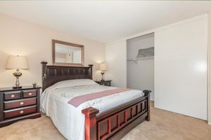 A cozy bedroom featuring a dark wooden bed with a white quilt and pink throw, two matching bedside lamps on wooden nightstands, and a mirror above the bed. A closet with sliding white doors is visible in the background, and the room has neutral-colored walls and carpet.