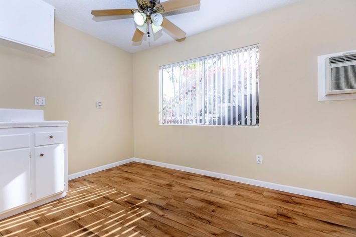 A small, empty room with light-colored walls and wooden flooring. It features a window with vertical blinds allowing sunlight to filter in, a ceiling fan, and a wall-mounted air conditioning unit. A small white kitchen area is visible in the corner.