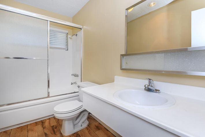 A bright bathroom featuring a white sink and countertop, a toilet, and a glass-enclosed shower. The walls are painted a light beige, and there's a large mirror above the sink. Light wood flooring complements the modern design. Natural light filters through a small window.