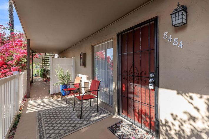 A cozy entryway featuring a red door with a decorative grille, flanked by two red chairs on a patterned rug. Bougainvillea plants add color outside, and a small potted plant sits nearby. The corridor is well-lit, with a lantern above the door, and a white railing leading to a pathway.