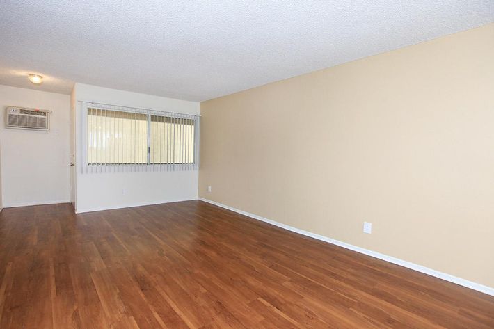 A spacious, unfurnished interior of a room featuring light-colored walls, wood laminate flooring, and a large window with vertical blinds letting in natural light. An air conditioning unit is mounted on the wall. The overall atmosphere is bright and inviting, ideal for personalization.