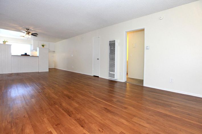 A spacious, light-filled living area featuring hardwood floors, white walls, and a ceiling fan. To the left, an open kitchen area is partially visible, with a small bar counter. A doorway leads to another room, while a wall-mounted heater is located on the right. The overall atmosphere is clean and inviting.