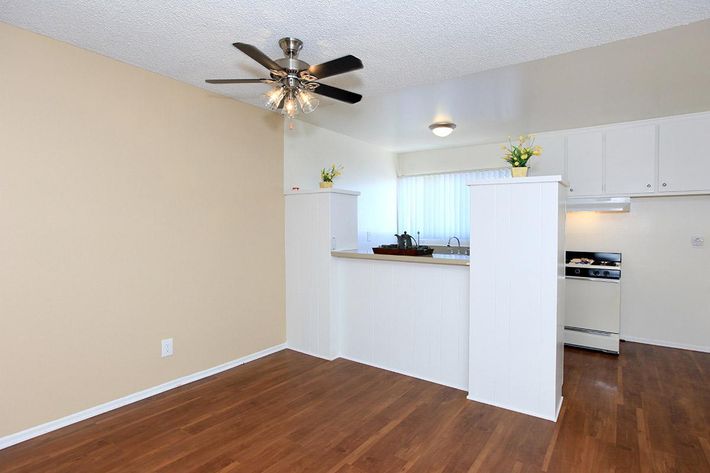 A well-lit kitchen and dining area featuring light beige walls, a wooden floor, and a ceiling fan. The kitchen includes white cabinetry and a gas stove, with a small bar area separated by two white partitions. Natural light filters in from a window, enhancing the warm atmosphere of the space.